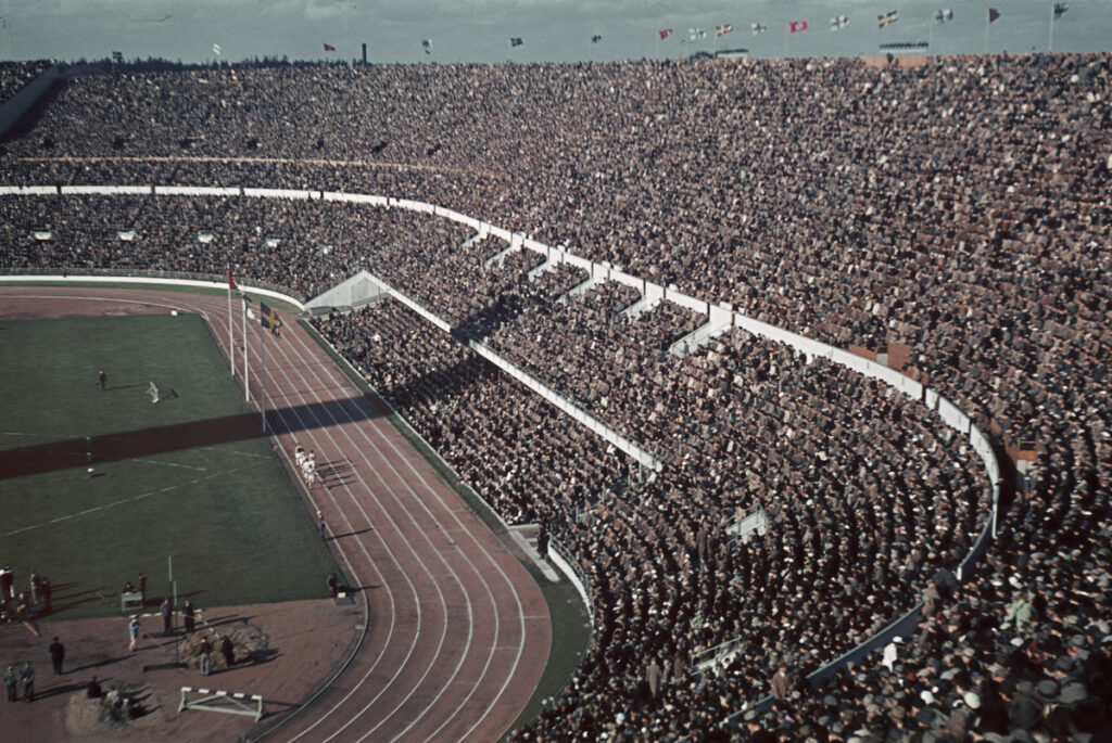 Kuvassa Helsingin olympiastadion, jonka katsomo on täynnä ihmisiä. Saloissa liehuvat Suomen, Ruotsin ja Saksan liput vuonna 1940.