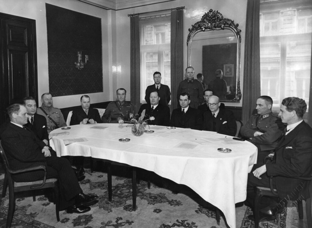 Kuvassa pöydän ääressä istuu yksitoista vakavailmeistä miestä puvuissa ja asepuvuissa. Takana seisoo kaksi miestä.
